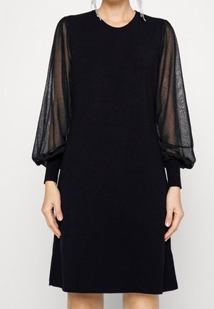 Robe noire longueur genoux avec manches longues bouffantes en tulle transparent et poignets ajustés portée par une personne avec des boucles d’oreilles en cristal pendantes.