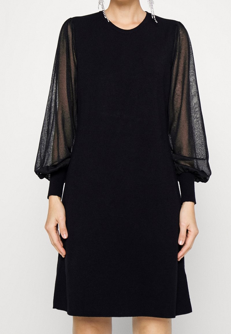 Robe noire longueur genoux avec manches longues bouffantes en tulle transparent et poignets ajustés portée par une personne avec des boucles d’oreilles en cristal pendantes.