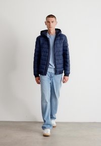 Jūras spēku pūkains mētelis ar kapuci, ar quilted tekstūru un priekšējo rāvējslēdzēju. Uzvilkts virs gaiši zila treniņjaka ar brīva piegriezuma gaišajām džinsām.