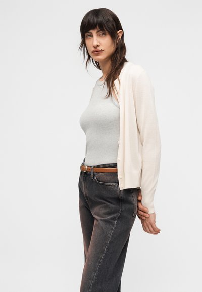 Νεαρή γυναίκα με σκούρα μαλλιά, που φοράει αμάνικο ανοιχτό γκρι τοπ, μπεζ πουλόβερ, μαύρα τζιν και καφέ ζώνη, πόζαρε μπροστά σε απλό λευκό φόντο.