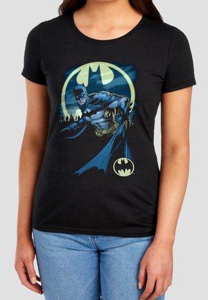 Frau trägt ein schwarzes T-Shirt mit Batman in Kampfpose mit einem leuchtenden Fledermaussymbol und Skyline der Stadt im Hintergrund.