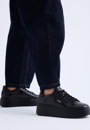 Persona que lleva vaqueros azul oscuro y zapatillas negras con plataforma con la marca "exe" sobre un fondo blanco.