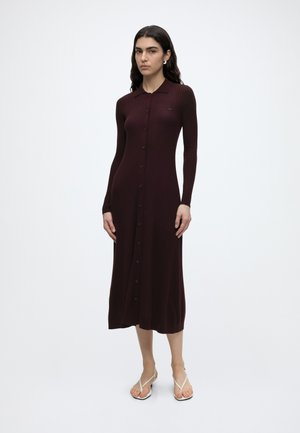 Mujer con un vestido midi acanalado, de manga larga, con botones, color burdeos oscuro, y cuello, de pie sobre un fondo blanco liso.