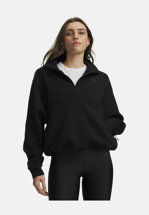 Vrouw draagt een zwart sweatshirt met kwart-rits over een wit shirt en zwarte leggings, staand met één hand in haar zak tegen een effen achtergrond.