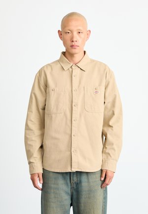 Light beige, long-sleeve button-up shirt with two chest pockets, stitched detailing, and a subtle logo patch. Smooth fabric texture.