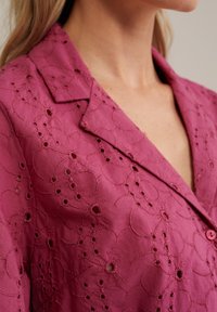 Rosa broderad blus med krage, med ett blommönster och små utskärningar, som visar en texturerad yta och knappstängning.