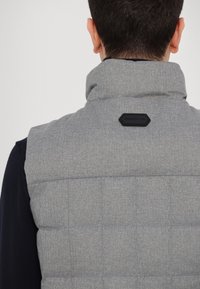 Gilet imbottito grigio con collo alto e motivo a rombi rettangolari. Presenta una toppa nera con logo sul retro e un maglione blu scuro sotto.