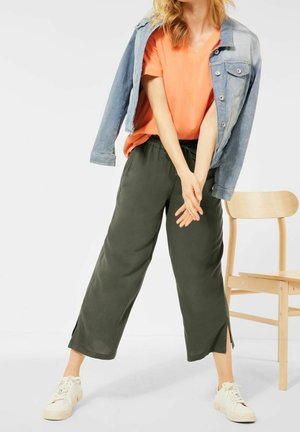 Personne portant une chemise orange, un pantalon large noir avec des fentes latérales, des baskets blanches, et une veste en jean drapée sur les épaules à côté d'une chaise en bois.