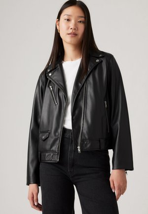 Schwarze Fake-Leder-Bikerjacke mit silbernen Reißverschlüssen, asymmetrischem Kragen und Vorderaschen. Kombiniert mit einem weißen Oberteil und schwarzen Jeans.