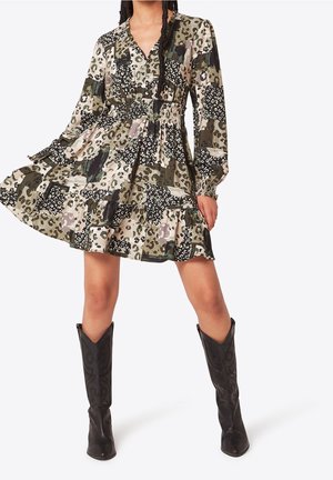 Femme portant une robe patchwork à imprimé animalier avec manches longues et taille élastique, associée à des bottes western noires montant jusqu'au genou.