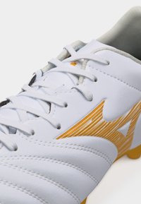 Κοντινό πλάνο λευκού ποδοσφαιρικού παπουτσιού με κίτρινο λογότυπο και κορδόνια, που δείχνει ραμμένο δέρμα και επενδυμένο κολάρο.