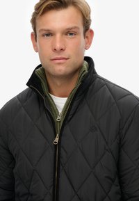 Chaqueta acolchada negra con un cuello interior de forro polar verde y cierre de cremallera. Presenta un pequeño logo en el pecho y costuras diagonales.