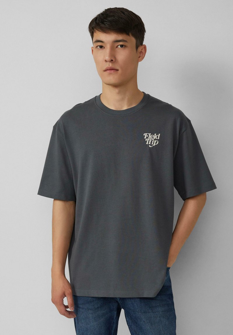 Jonge man met kort zwart haar, gekleed in een oversized donkergrijs T-shirt met de tekst "Field Trip" en een spijkerbroek, staande tegen een effen muur.
