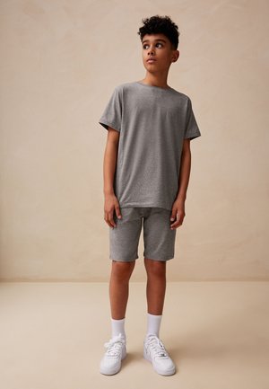 Nastolatek stojący w pomieszczeniu, ubrany w szarą koszulkę, pasujące spodenki, białe skarpetki i białe trampki, patrzący lekko w bok.