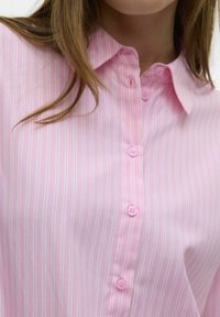 Lichtroze blouse met verticale witte strepen, knoopsluiting aan de voorkant met bijpassende roze knopen, en een klassiek kraagontwerp. Zachte stoftextuur.