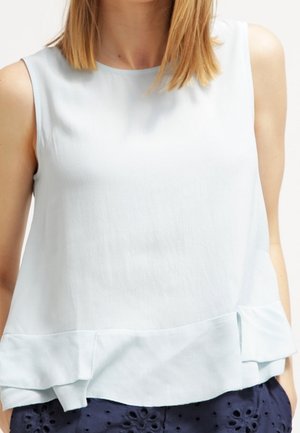 Kvinde iført en ærmeløs lyseblå bluse med flæsekant, kombineret med mørkeblå shorts med øjenvævning.