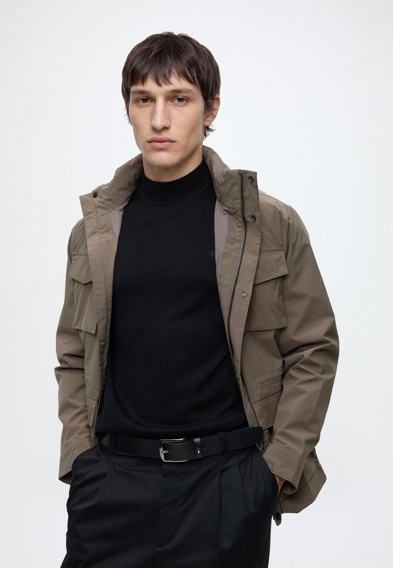 Young man with short dark hair wearing a tan jacket over a black turtleneck, black pants, and a black belt against a plain background.