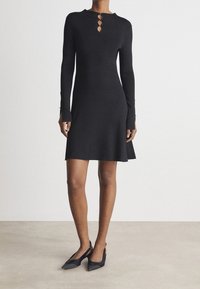 Robe noire en maille à manches longues avec un col haut et trois boutons en forme de trou de serrure à l'arrière ; corsage ajusté et ourlet évasé. Portée avec des talons noirs.