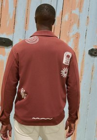 Maroonsweater med lange ærmer, prydet med hvide broderede motiver, herunder en spiral, blade og linjer på ryggen. Glat tekstur.
