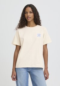 Lahka bež bombažna majica s kratkimi rokavi, z majhno modro-rdečo cvetlično grafiko na levi strani prsnega koša. Ravna obroba, sproščen kroj.