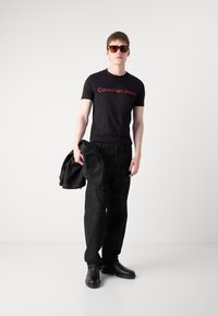 Mann med svart Calvin Klein Jeans t-skjorte, mørke jeans, svarte sko, solbriller, holder en mørk jakke mot en ensfarget lys bakgrunn.