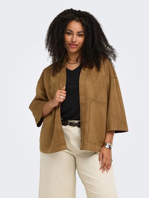 Kvinde med krøllet hår iført brun åben jakke foran, sort top, beige bukser, sort bælte og sølvur mod ensfarvet baggrund.