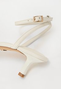 Vita läderklackad sandal med en smal ankelrem med ett guldspänne, en elegant tårem och en ljusbrun fyrkantig klack.