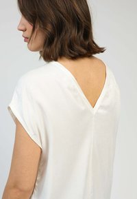 Blouse blanche à manches courtes avec un décolleté en V dans le dos. Texture lisse avec un éclat subtil. Le design présente une coupe ample et un style minimaliste.