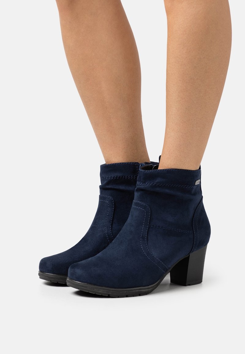 Jambes portant des bottines en daim bleu marine avec talons carrés et surpiqûres latérales, debout sur une surface blanche.
