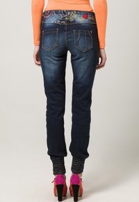 Nainen, jolla on tummansiniset farkut, joissa on kirjailua vyötäröllä, oranssi paita ja vaaleanpunaiset-oranssit korot, seisoo selkä kameraan päin.