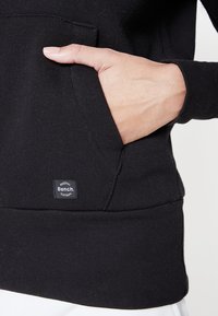 Schwarzer Hoodie mit Kängurutasche, ausgestattet mit gerippter Textur und einem kleinen Markenlabel an der unteren Vorderseite. Weicher Stoff, lässiges Design.