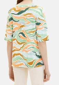 Femme portant une chemise ample à manches courtes avec un motif abstrait ondulé en vert, orange, blanc et marron, associée à un pantalon beige clair.