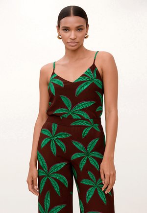 Mujer con top y pantalones marrones sin mangas con grandes patrones de hojas de palma verdes, pendientes de aro dorados y el cabello peinado hacia atrás.