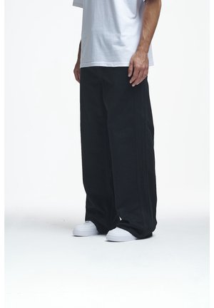 Personne portant un pantalon noir ample, des baskets blanches et un t-shirt blanc, debout sur un sol blanc devant un fond uni.