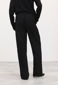 Pantalon noir à jambe large avec une ceinture élastique, doté de deux poches arrière et d'une texture de tissu lisse, associé à des chaussures noires.