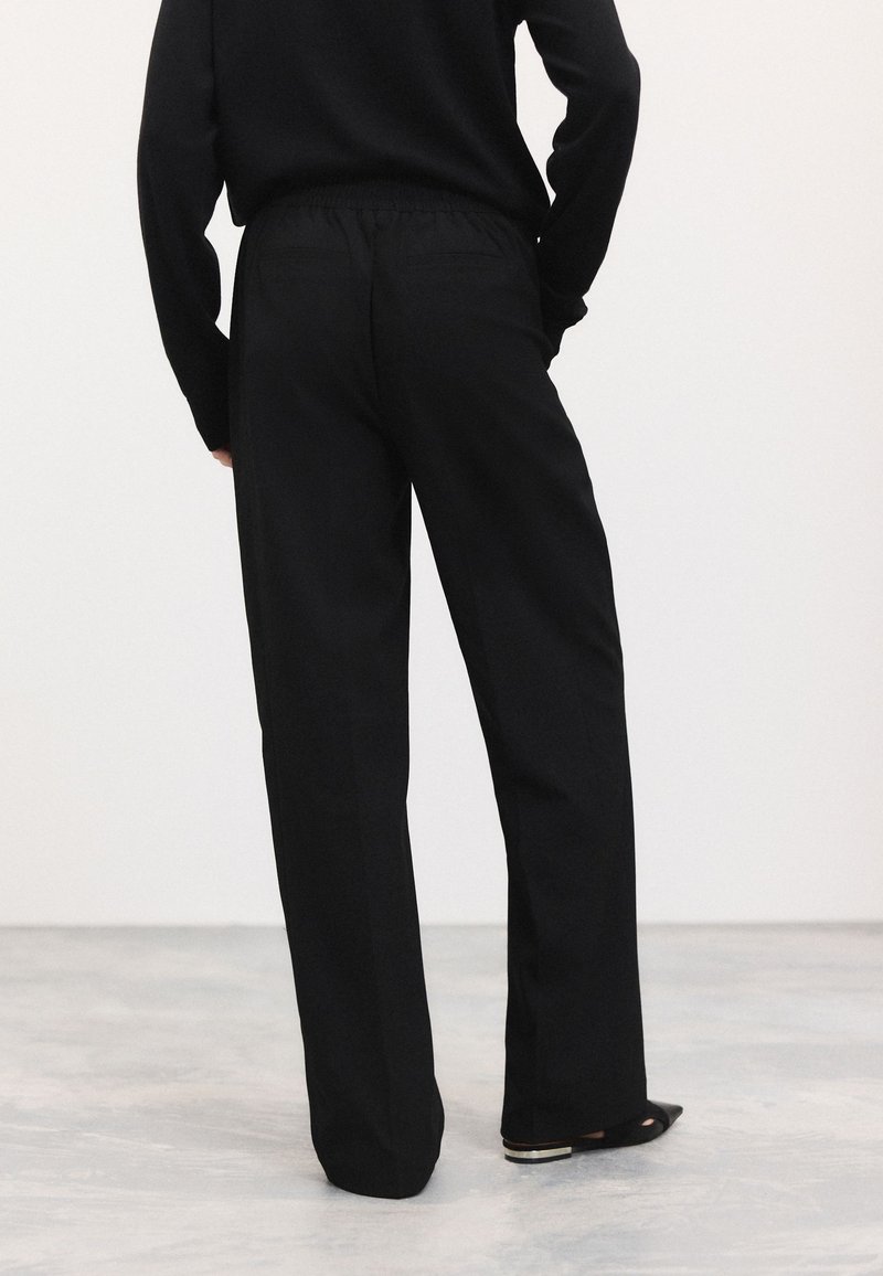 Pantalon noir à jambe large avec une ceinture élastique, doté de deux poches arrière et d'une texture de tissu lisse, associé à des chaussures noires.