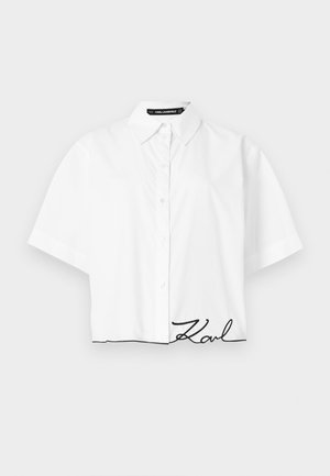 Λευκό πουκάμισο με κουμπιά και κοντά μανίκια, με γιακά και μαύρη κεντημένη υπογραφή "Karl" κοντά στο κάτω τελείωμα σε απλό φόντο.