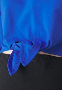 Mėlynas medvilninis marškinėliai su užrišta hemline detale nugarėje. Juodas elastinis juostos kraštas matomas apačioje. Lygi tekstūra.