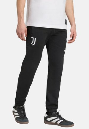 Man draagt zwarte joggingbroek met Juventus-logo en zwarte sneakers met witte strepen, staand tegen een witte achtergrond.