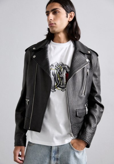 Black leather motorcycle jacket with textured finish, silver zippers, shoulder epaulets, worn over a white graphic t-shirt and denim jeans.
