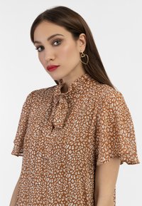 Brun bluse med et lyst dyreprintdesign, korte voluminøse ærmer og en sløjfe-halsudskæring. Gyldne hjerteformede øreringe og en glat tekstur.