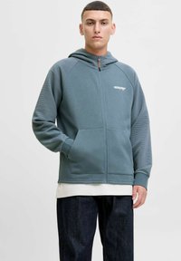 Sweat à zip couleur sarcelle avec des textures côtel�ées aux épaules, des poches avant et un détail de logo blanc. Porté par-dessus une chemise blanche à couches.