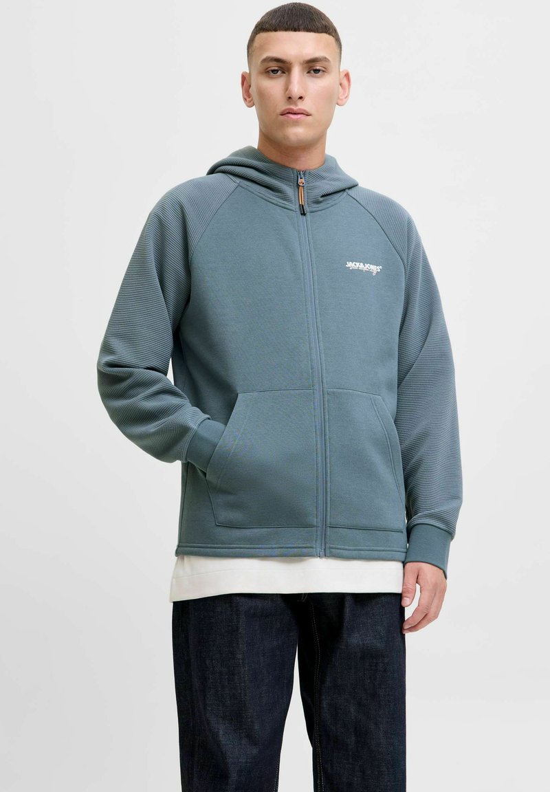 Sweat à zip couleur sarcelle avec des textures côtelées aux épaules, des poches avant et un détail de logo blanc. Porté par-dessus une chemise blanche à couches.