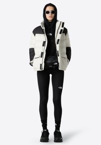 Puffjakke i creme med sorte detaljer, lynlåslukning, ledsaget af en sort hættejakke, leggings og sorte sneakers. Solbriller tilføjet.