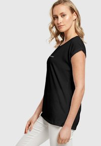 Sort bomulds t-shirt med rullede korte ærmer, rund halsudskæring og logo detalje foran. Båret med lysfarvede slim-fit bukser.