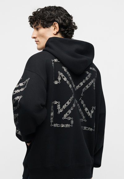 Jauns vīrietis ar cirtainiem matiem, valkājot melnu kapuci ar lielu baltu sarežģītu ķēdes rakstu uz muguras un piedurknēm.