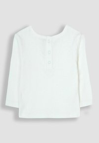 Maglia bianca a maniche lunghe realizzata in morbido tessuto. Collo rotondo con quattro bottoni sul retro e intricati dettagli a occhielli sulle spalle.