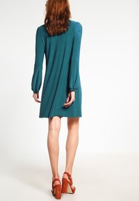 Teal, långärmad klänning med avslappnad passform, rynkade ärmslut och en svängig nederkant. Bärs med bruna platåsandaler.