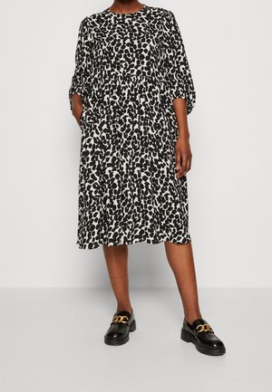 Vrouw in een zwart-witte midi-jurk met dierenprint en geplooide driekwartsmouwen, draagt zwarte loafers met een gouden kettingdetail, staand tegen een witte muur.