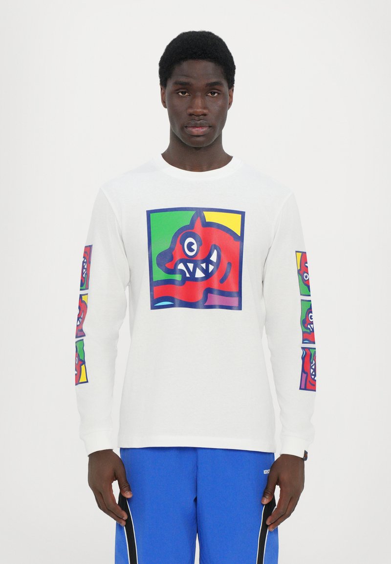 Langärmliges weißes T-Shirt mit einem bunten Grafikdruck eines roten cartoonartigen Gesichts auf der Vorderseite und kleineren Versionen an den Ärmeln.